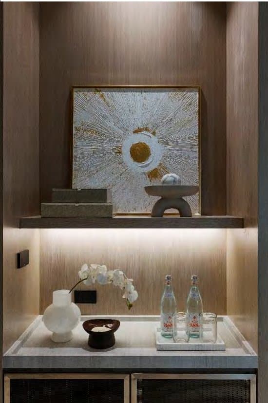 Warm backlit niche framing a gold-flecked sunburst artwork, white orchid, textured vase and sparkling water on a stone tray.