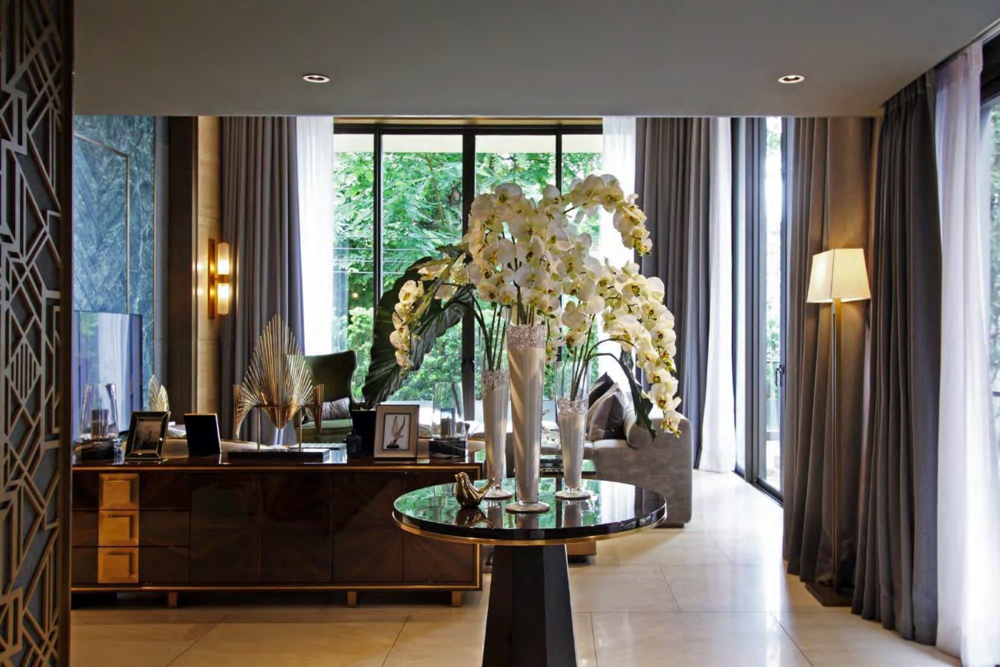 Living salon with a walnut console, black glass table and towering white orchids set before floor-to-ceiling windows onto lush greenery