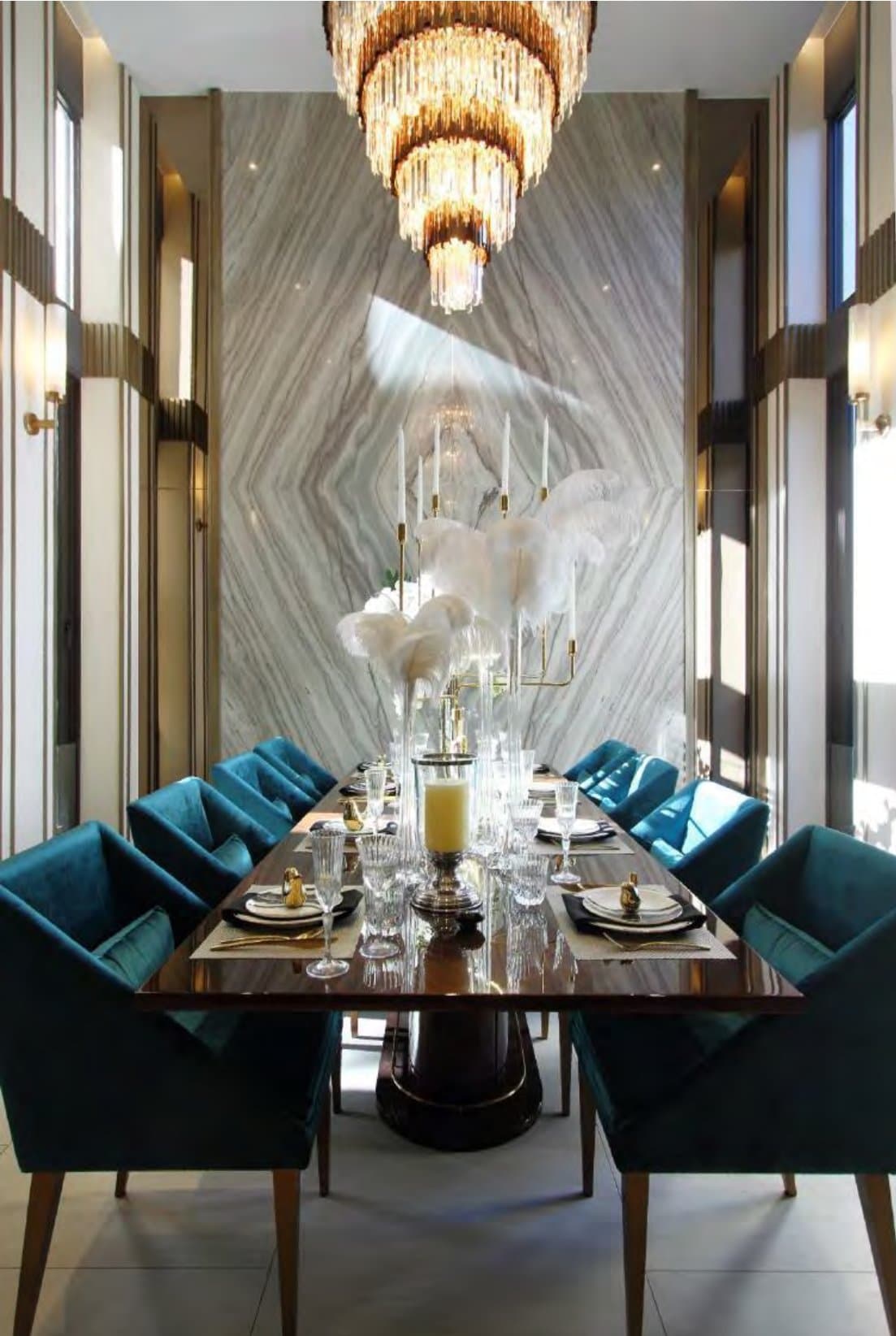Formal dining room with a tiered crystal-and-brass chandelier above teal velvet chairs, a timber table and book-matched marble wall
