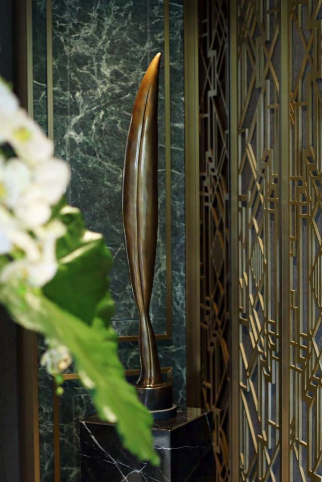 Bronze leaf-form sculpture on a black marble plinth against green marble and a brass deco fretwork screen, white orchids nearby