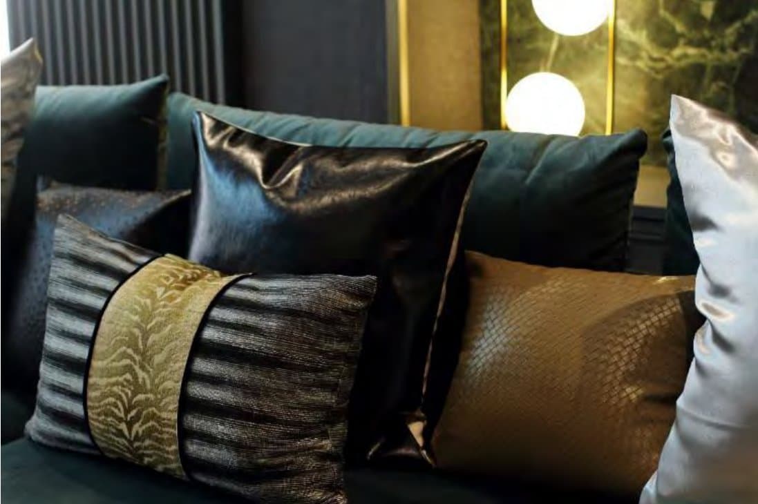 Layered cushions in teal velvet, black leather, gold and silver satin against backlit green marble and a glowing globe lamp