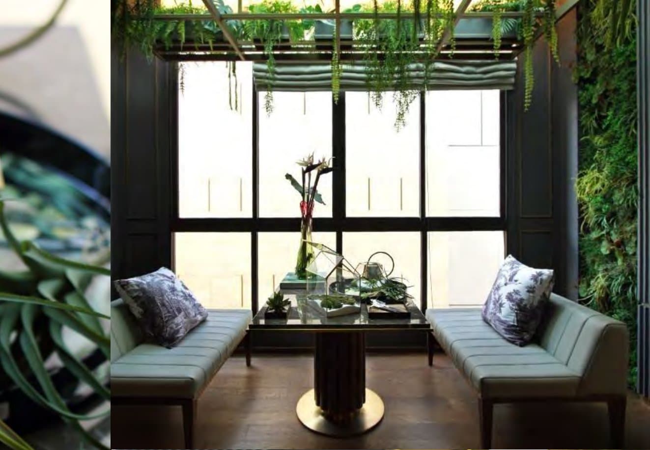 Garden room with grey banquettes flanking a brass-footed marble table, black steel glazing and a living green wall of ferns
