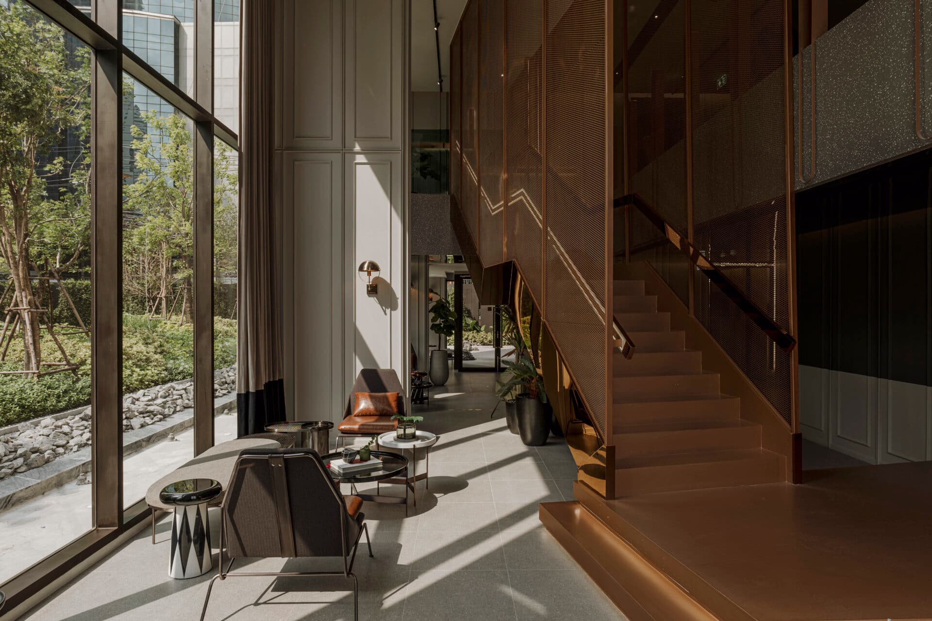 Double-height lobby lounge with floor-to-ceiling windows onto a garden, bronze mesh staircase, armchairs and round coffee tables