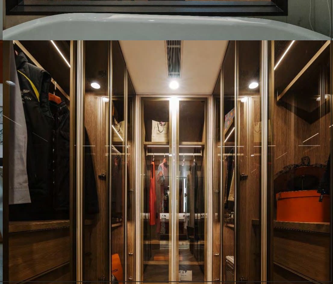 Walk-in wardrobe lined with glass-front timber cabinets, warm downlights, hanging jackets, a red dress and displayed accessories