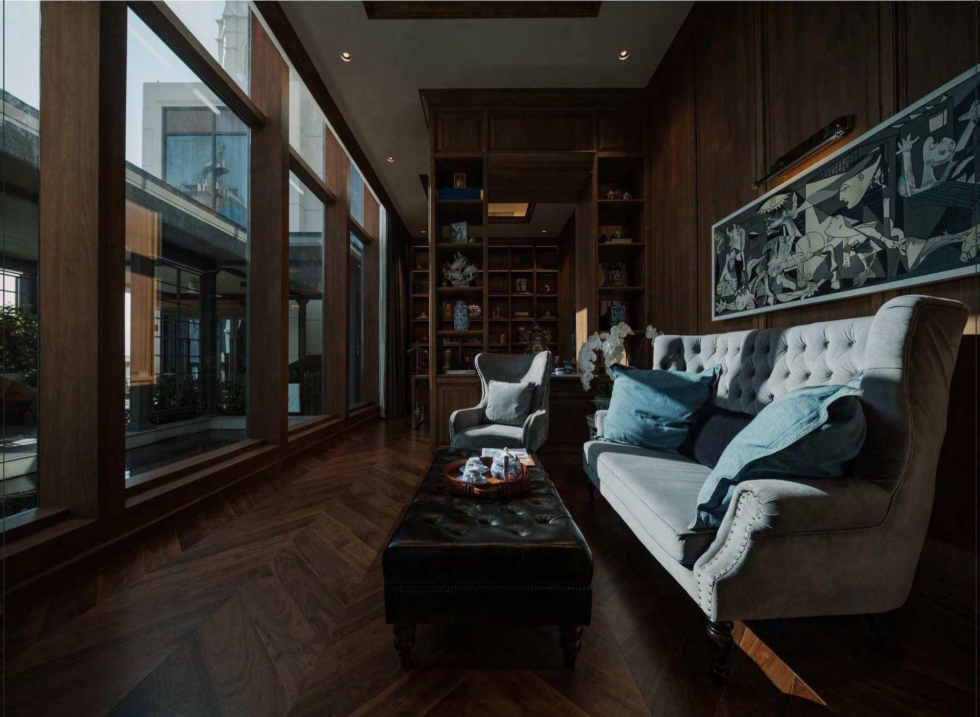 Panelled library with a grey velvet sofa, buttoned leather ottoman and monochrome mural, tall windows framing a neighbouring tower