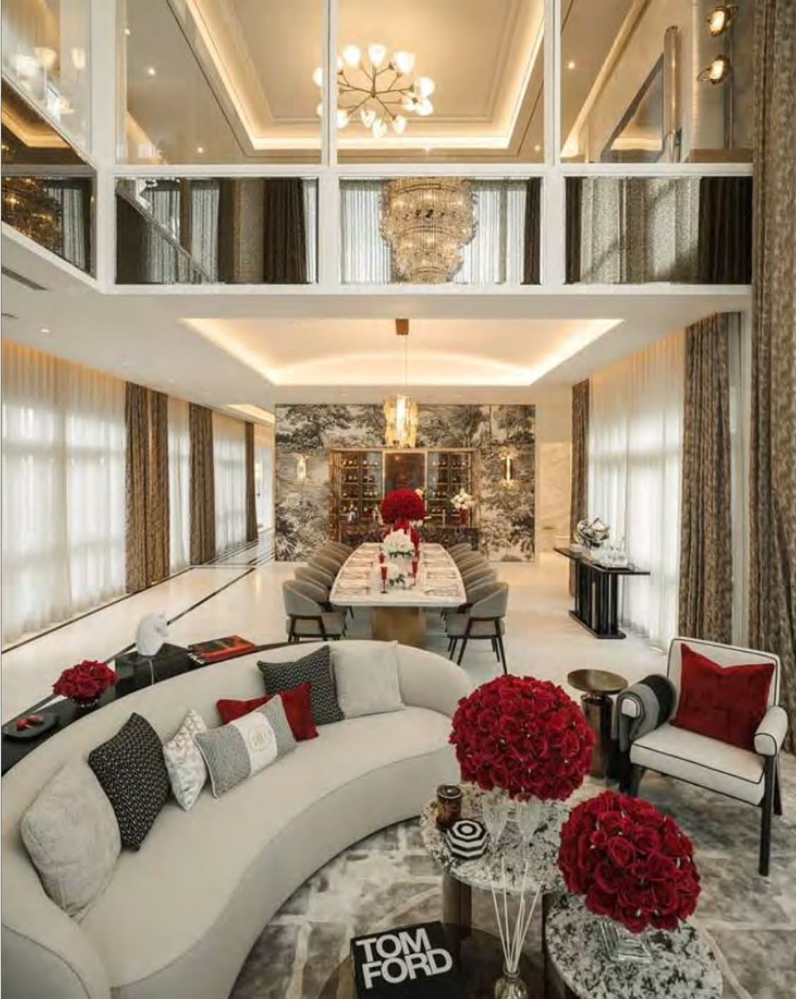 Double-height reception with a curved cream sofa, domed red rose arrangements, gold sputnik and crystal chandeliers, and a formal dining table beyond.
