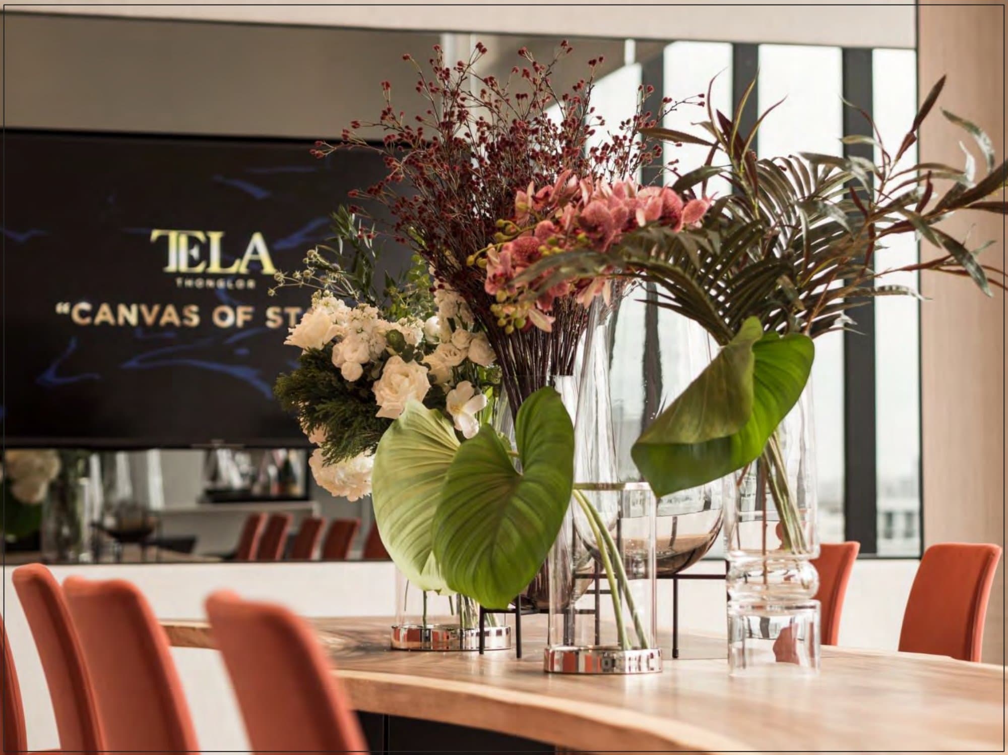 Sculptural floral arrangement of orchids, berries and broad tropical leaves crowns a timber table before a backlit 'Canvas of Style' screen and coral chairs