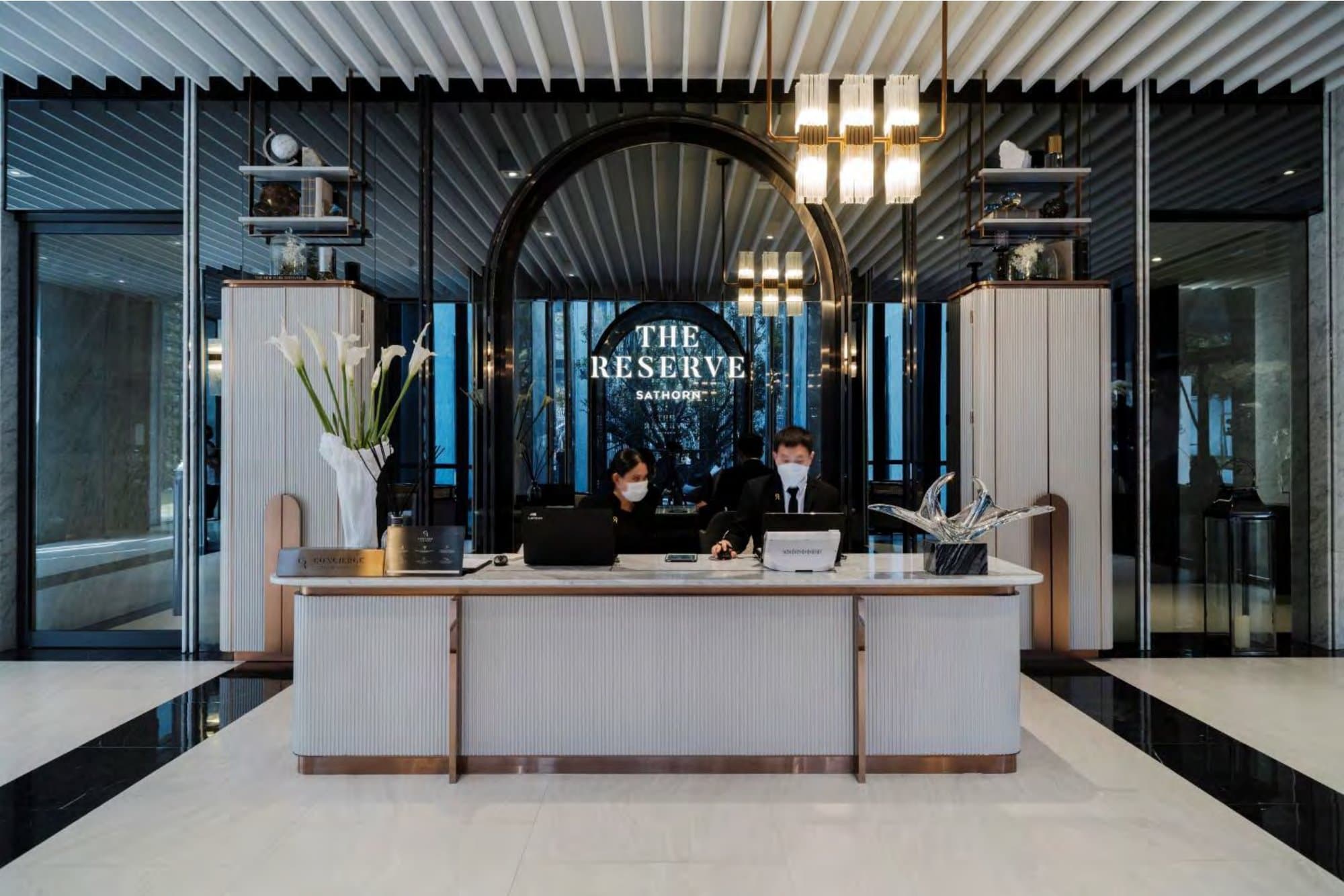 Condominium concierge lobby with a black arch, illuminated Reserve wordmark, fluted crystal pendants, pale marble desk and white calla lilies